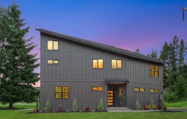 Modern house with large windows and a sloped roof, surrounded by greenery at sunset.