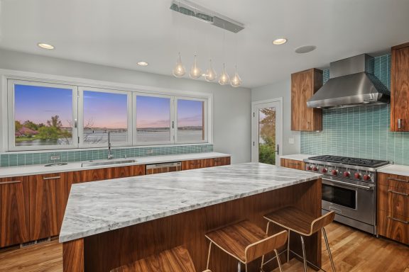 2019 Modern kitchen with marble countertops, wooden cabinets, and large windows showcasing a sunset view.